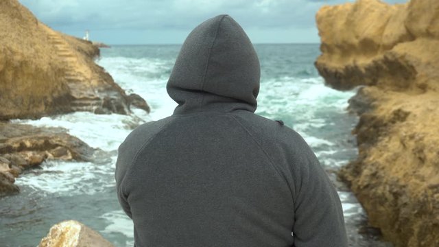 A man in a hood stands before a cliff in the sea. The sea among the rocks. A man stands with his back. Storm on the Mediterranean Sea.