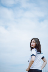 pretty asian lady with short straight hair in Japanese student uniform posing standing turned back in the park, turning to look and smile a little at camera with a beautiful sky background