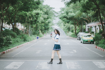 pretty asian girl in Japanese student uniform with short dark hair holding an umbrella and walking across the street in the middle of somewhere with a lot of trees along the street