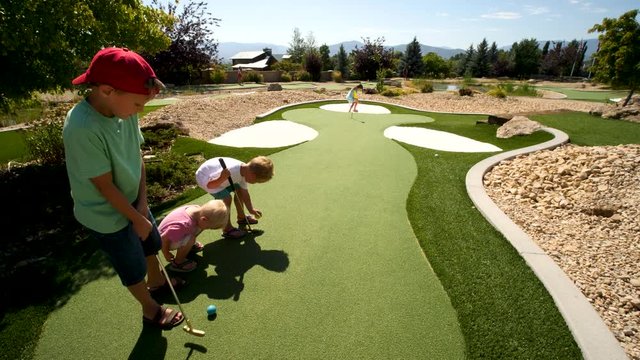 Brothers are standing near their sister as he tees off. Close up hand held video showing four siblings at mini golfing yard and sister drives the ball. You can see the trees at the background in this.