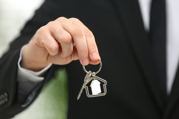 Real estate agent holding house key with trinket on blurred background, closeup