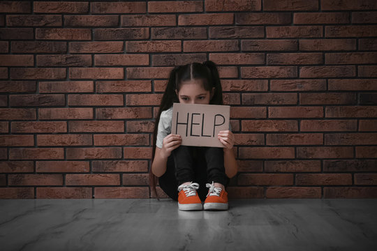 Sad little girl with sign HELP on floor near brick wall. Child in danger