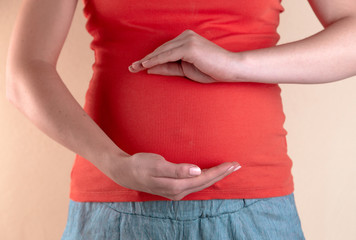A close-up view of the belly of a pregnant woman in a red T-shirt that holds her hands shape health care digestion on body closeup belly. The concept of love and care during pregnancy.