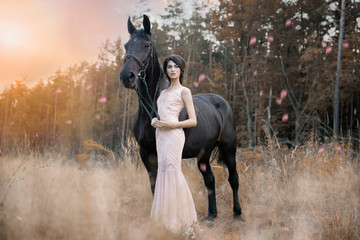 Fototapeta na wymiar Beautiful art picture with a black crow mare and a girl in a pink dress at sunset in a whirlwind of pink petals