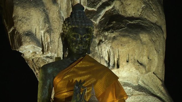 Close up of a Buddha image inside a cave, well illuminated, wearing an orange sash, and in front of a large boulder