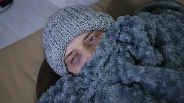 Covered with grey plaid young caucasian man in hat and scarf freezing feeling cold at home, ill sick male having fever flu influenza temperature symptoms wrapped in blanket shivering indoors.