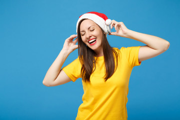 Cheerful young woman Santa girl in Christmas hat posing isolated on blue background. New Year 2020 celebration holiday concept. Mock up copy space. Listen music with headphones, keeping eyes closed.