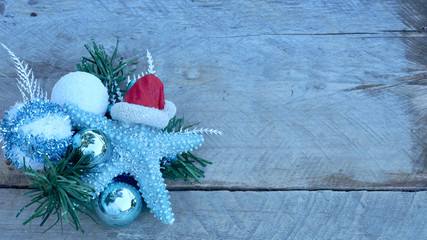 turquoise starfish with red Santa Claus hat laying on a pine branch with christmas bulbs on a natural wood background with copy space