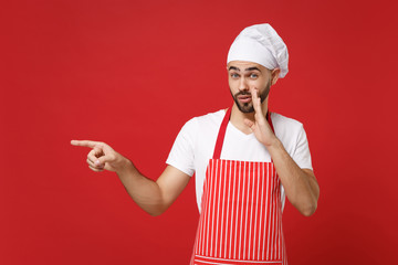 Secret chef cook baker man in apron toque chefs hat posing isolated on red background. Cooking food concept. Mock up copy space. Whispers gossip, tells secret with hand gesture, pointing finger aside.