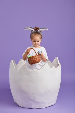 Happy Child Girl In Costume Easter Bunny Rabbit With Ears And A Basket With Eggs. Little Girl In A Big Chicken Egg On A Purple Background