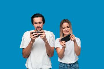 Beautiful couple wearing white t-shirts standing isolated over blue background, playing games on mobile phone