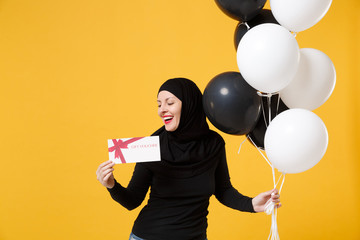 Arabian muslim woman in hijab celebrating hold voucher black white air balloons isolated on yellow background studio portrait. Birthday holiday people religious lifestyle concept. Mock up copy space.