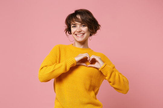 Smiling young brunette woman girl in yellow sweater posing isolated on pastel pink background in studio. People lifestyle concept. Mock up copy space. Showing shape heart with hands, heart-shape sign.