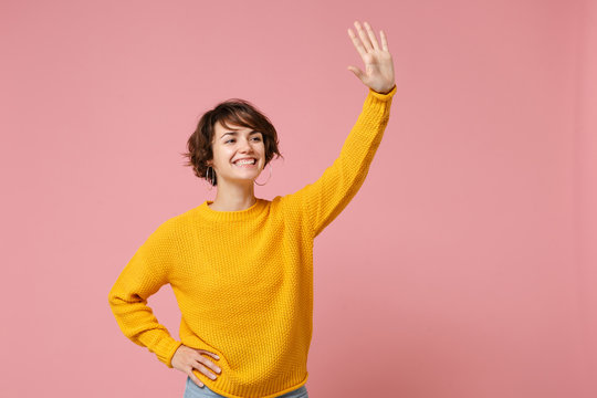 Pleasant young brunette woman girl in yellow sweater posing isolated on pink background studio portrait. People lifestyle concept. Mock up copy space. Waving and greeting with hand as notices someone.