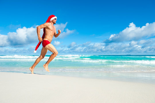Athletic Santa with long hat running in red sport briefs on the shore of a tropical beach in a Christmas workout routine