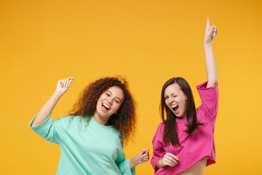 Two cheerful women friends european african american girls in pink green clothes posing isolated on yellow background. People lifestyle concept. Mock up copy space. Having fun dancing rising hands up.