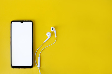 Smartphone with blank white screen connects to earphones on yellow background - top view. Earphone...