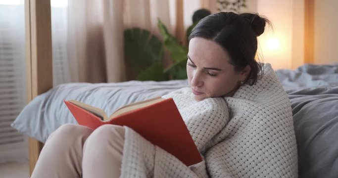 Young woman reading book wrapped in a warm blanket at home