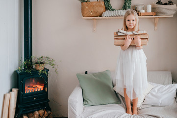 Beautiful little girl in white dress standing on bed with woods in her hands in chalet style room...