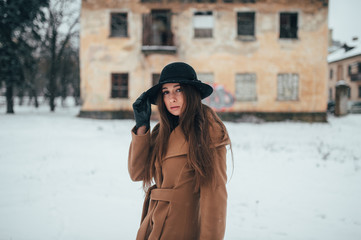 Young stylish girl in brown coat and black hat posing outdoor in winter against abanoned house.