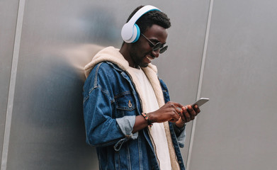 Cool modern smiling african man in wireless headphones listening to music using phone on city...