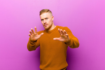 young blonde man wearing a jumper feeling terrified, backing off and screaming in horror and panic,...