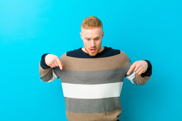 young blonde man feeling shocked, open-mouthed and amazed, looking and pointing downwards in...