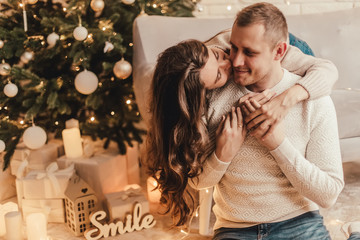 Fototapeta premium Beautiful young couple kisses sitting on the floor and celebrating new year in front of Christmas tree.