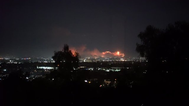 Forest fire on mountain range above residential neighborhood at night as planes dump water and smoke spreads helicopters fly around with search lights.