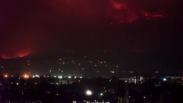 Forest fire on mountain range above residential neighborhood at night as planes dump water and smoke spreads helicopters fly around with search lights.
