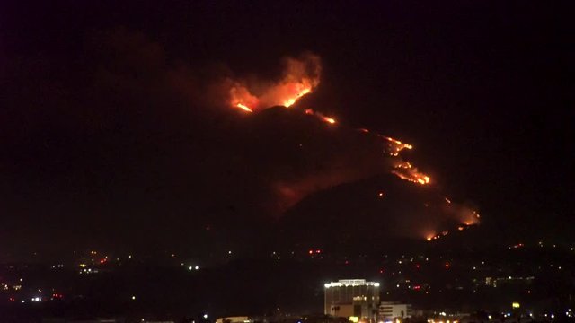 Forest fire on mountain range above residential neighborhood at night as planes dump water and smoke spreads helicopters fly around with search lights.