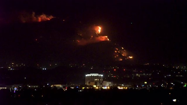 Forest fire on mountain range above residential neighborhood at night as planes dump water and smoke spreads helicopters fly around with search lights.