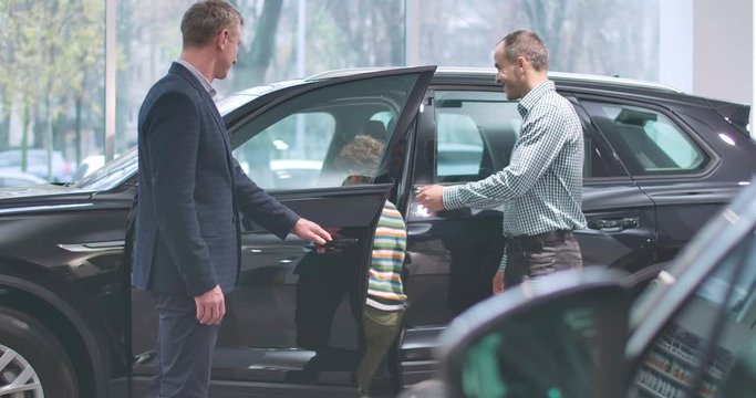 Male Caucasian car dealer talking to adult man and his son. Trader opening car door and little boy sitting into salon. Cheerful child choosing new automobile with father. Cinema 4k footage ProRes HQ.