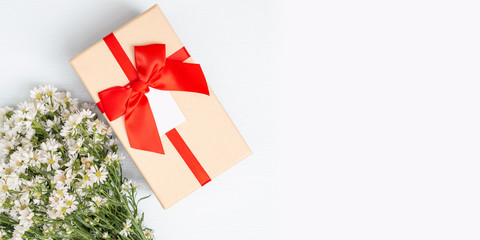 Gift box with red ribbon and white cutter flower on left side of white wooden background. Top view. Gift time. Copy space.