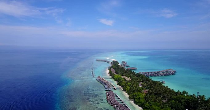 Idyllic tropical island with bungalows over shallow clear water of lagoon surrounded by large coral reef continuing to deep blue ocean horizon in Maldives 