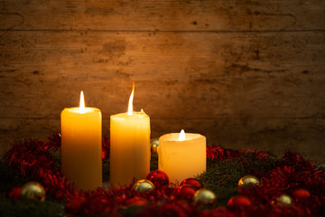 Obraz na płótnie Canvas The warmth of the Christmas concept: three candles lit on a light wooden table and a rustic setting with pine branches, red decoration and gold and red bright baubles with bokeh effect
