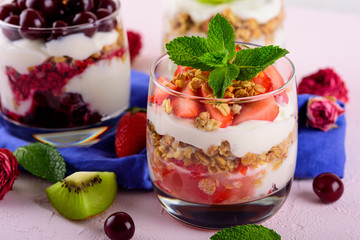 trifles with cherries, strawberries, whipped cream and granola in glass on blue napkin on light pink background