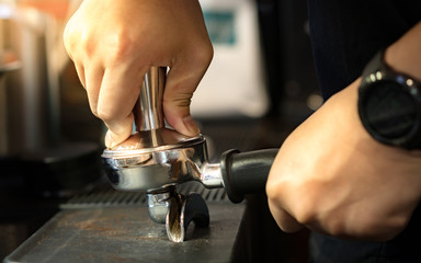 Closeup, barista's right hand hold coffee tamper and tamp arabica grounds in espresso double spout portafilter gripped by left on tamp mat before brewing with espresso machine. Practice makes perfect.