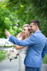Young beautiful couple of newlyweds bride and groom walk in a park with green trees on their wedding day and look in the mirror on the phone. Love and love story of wife and husband.