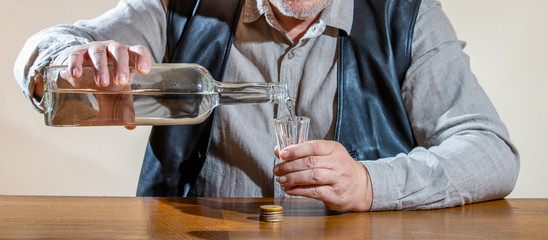 A man with a cap sits drunk at a table. A bottle of alcohol and a glass stand in front of him....