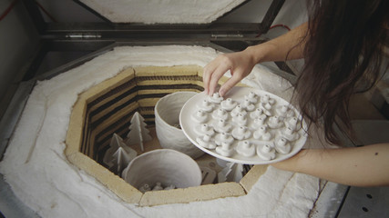 Closeup of asian female master potter puts small raw clay products into a kiln for firing....
