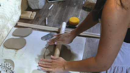 Top view of a woman potter wearing an apron preparing and rolling clay with a rollingpin. Female hands rolling out raw clay with wooden rolling pin