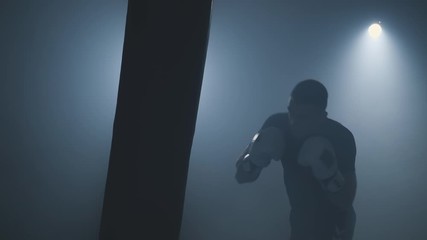 Strong athlete hits punching bag in smoky gym in slow motion. Boxer in gym. Young man training indoors. Sport concept. Sportsman boxing in dark studio. Low key. 4k, UHD