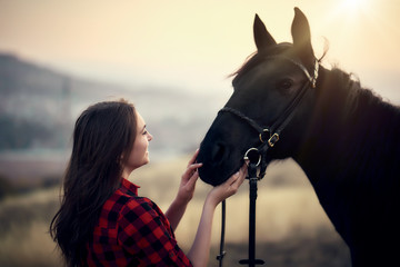 lovely young girl in a plaid shirt hugs a black horse at sunset in the mountains