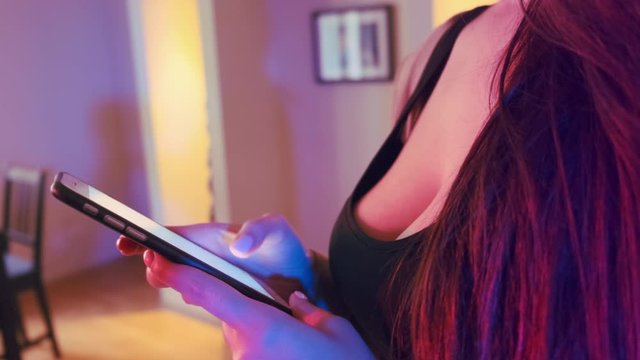Close up of sexy Caucasian young woman with big boobs and black decorative dress using smartphone at home. Neon lights interior. 10Bit ProRes Color Graded. Camera pan rotating left to right and forth.