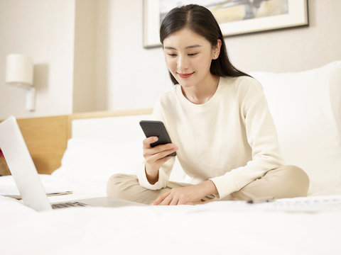 young asian woman using cellphone and laptop on bed