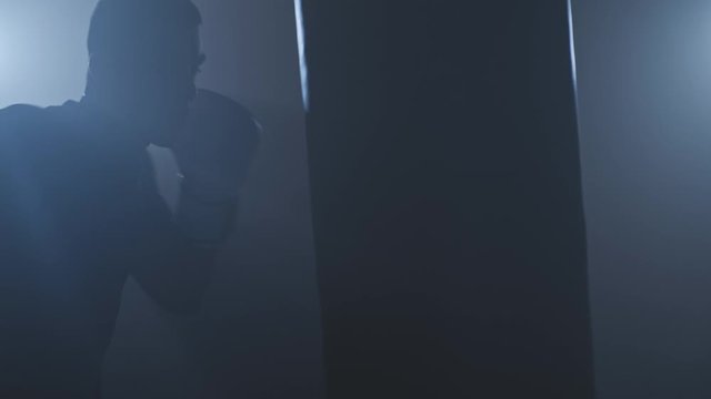 Strong athlete hits punching bag in smoky gym in slow motion. Close-up of boxer in gym. Young man training indoors. Sport concept. Silhouette of sportsman boxing in dark studio. Low key. 4k, UHD