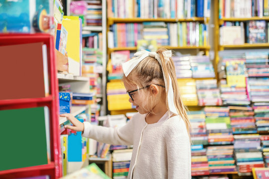 Side View Of Charming Caucasian Blond Girl With Ponytail And With Eyeglasses Standing In Bookstore And Choosing What To Read Next.