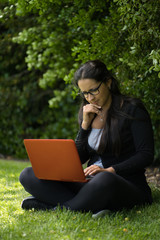 Brunette woman wearing glasses is sitting on the grass with her laptop. She is with her legs crossed and she is thoughtful. Vertical photography. Educational concept