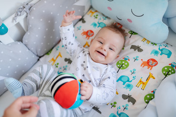 Top view of adorable Caucasian little baby boy with blue eyes lying in crib, catching stuffed ball and looking at camera.
